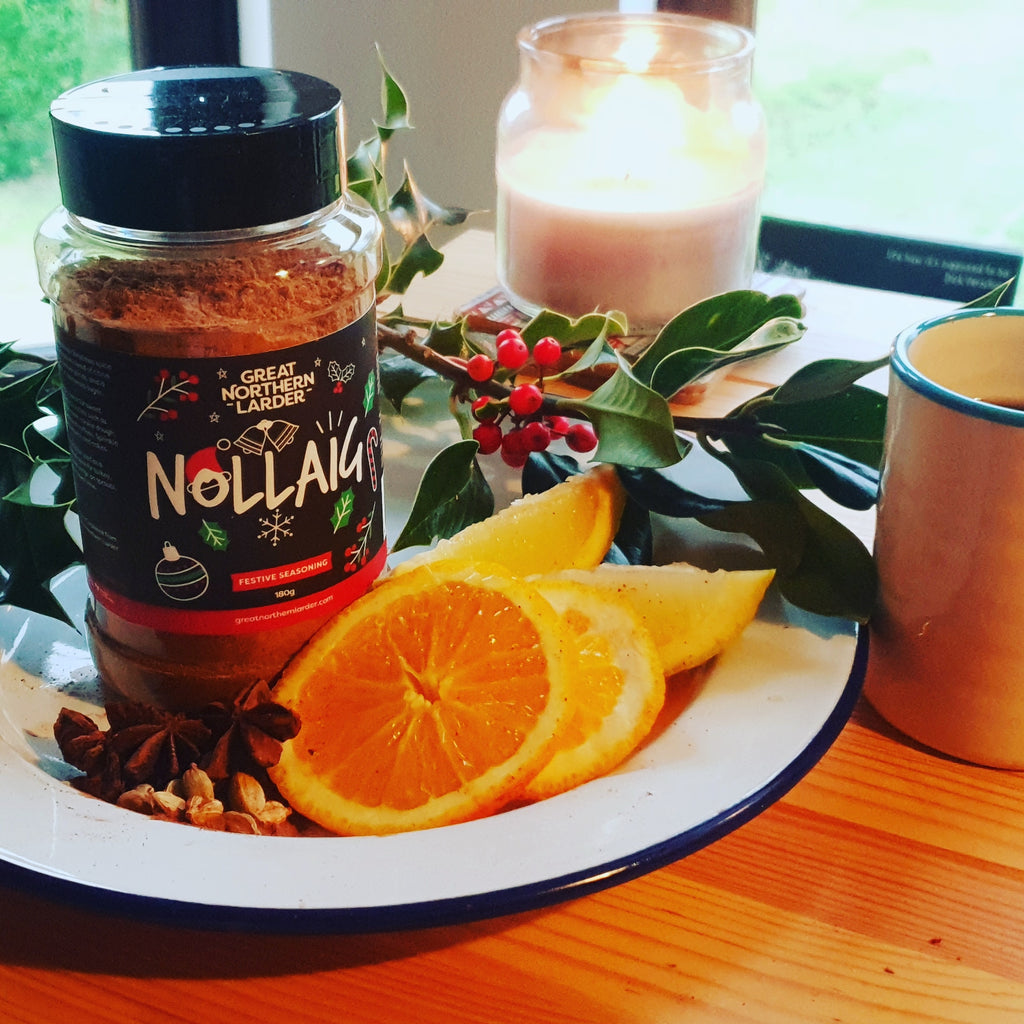 A photograph of Nollaig seasoning on a white enamel plate with orange slices, cloves, star anise and cardomom pods, surrounded by holly, berries, candles and a cup of mulled wine.